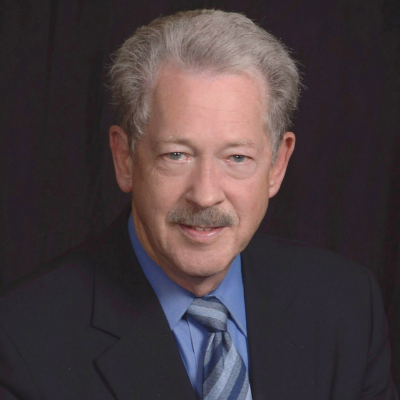 Thomas Risso's headshot. Tom is in a black suit with a blue shirt and gray and blue striped tie. He is in front of a plain black background and is smiling