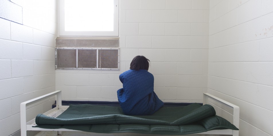 Photo of youth sitting on bed in empty solitary cell, back to camera.