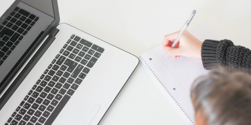 overhead view of someone taking written notes with a laptop on their desk.