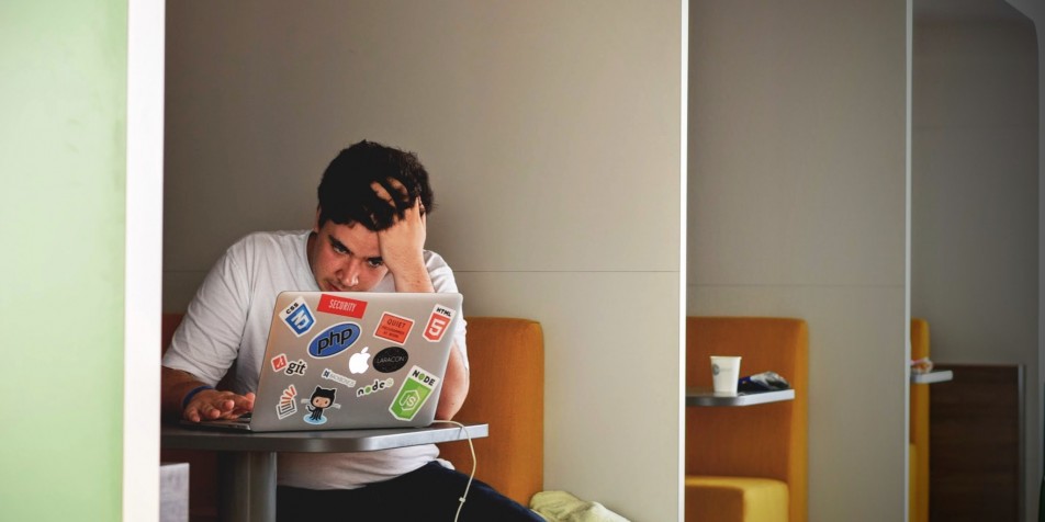 An image of a young man sitting at a table looking at his laptop.