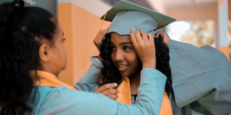 Two women in graduation gowns. One woman helps the other adjust her cap. 