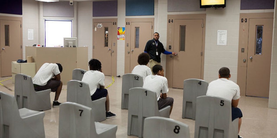 Four young men sit in numbered chairs in a detention facility.