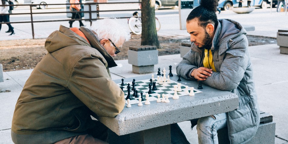 teen and older man playing chess outside in city. 