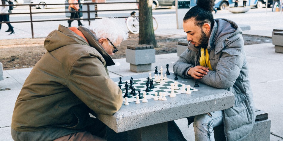 teen and older man playing chess outside in city.