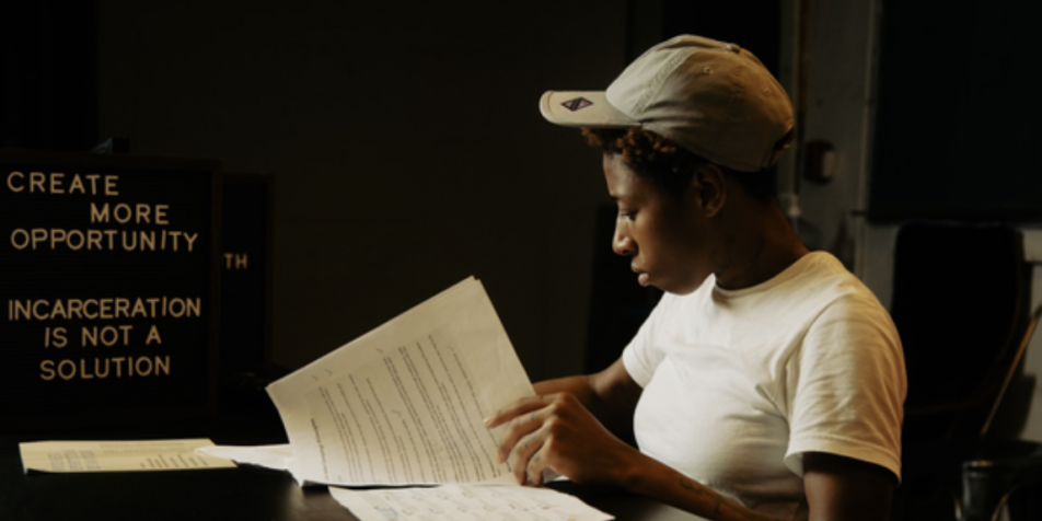 young person reading at a table in front of a sign that says "create more opportunity. incarceration is not a solution." 