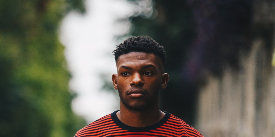 a young black man stands alone on a tree lined street in a red and black striped t shirt 