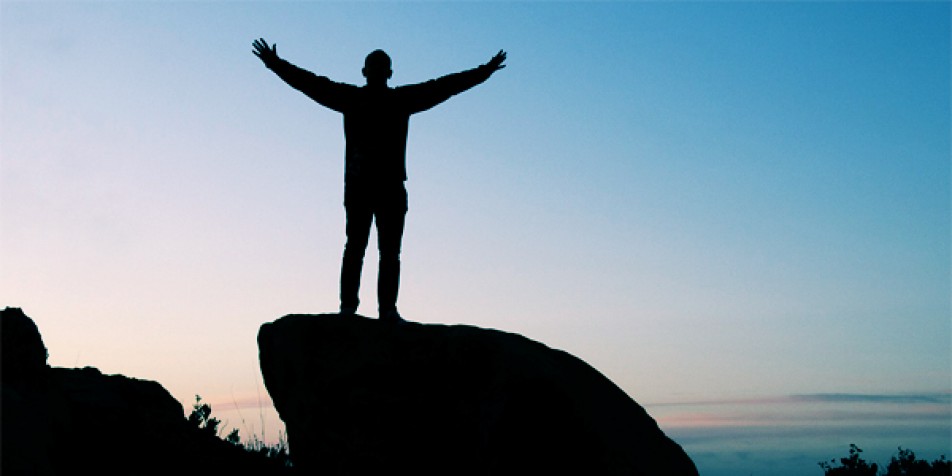 Silhouette of youth standing on rock holding arms up.