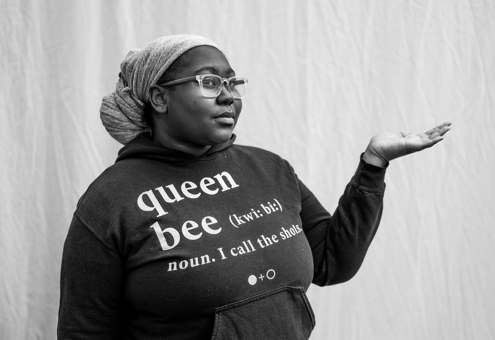 A person wearing glasses, a headwrap and a hooded sweatshirt with their hand extended, palm up